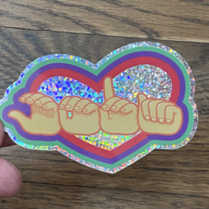 A die-cut sticker featuring American Sign Language (ASL) fingerspelling hands spelling out CODA (Child of a Deaf Adult) across a large heart background. The sticker has a wide, shimmering holographic glitter border, with a multi-colored red and green frame outlining the central heart.